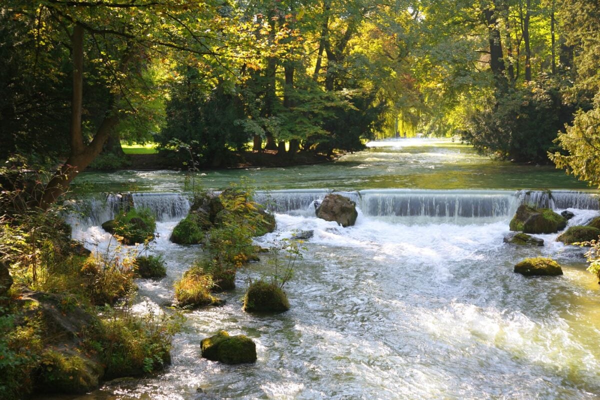 Small low waterfall on a rocky stream running through a sunlit wooded park.