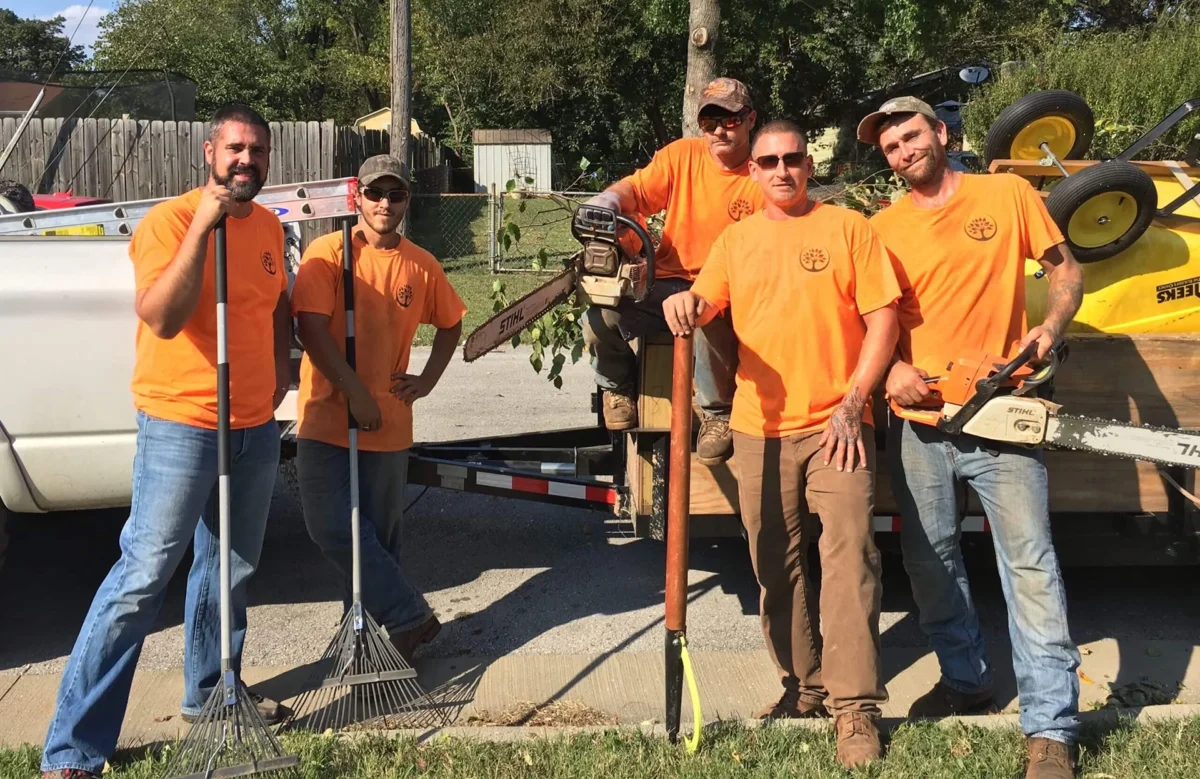 5-person crew posing with rakes/chainsaw and truck-and-trailer