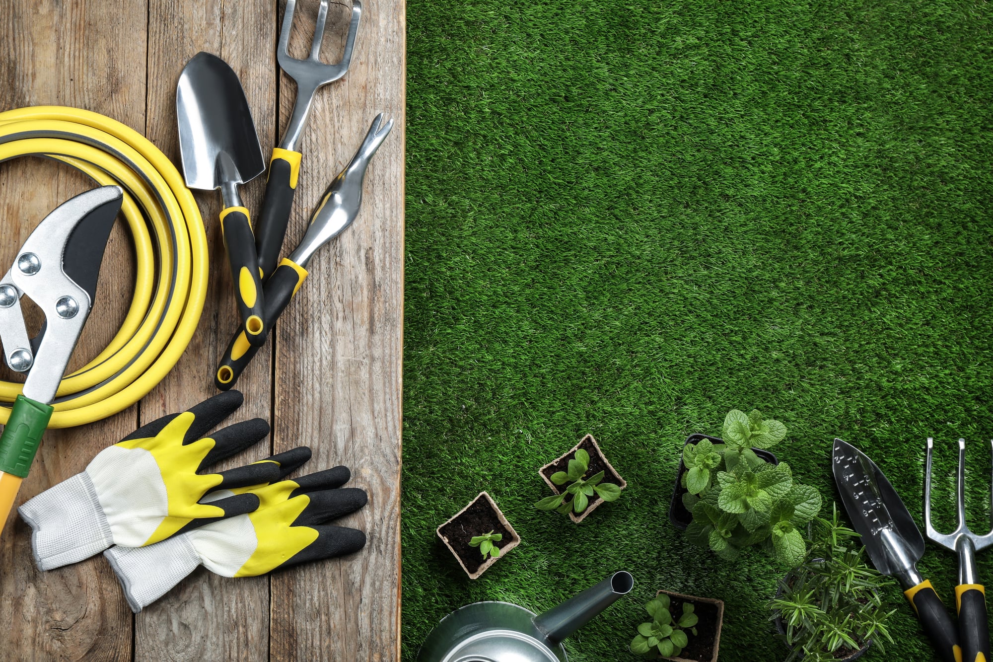 Gardening tools, work gloves and seedlings laid out on wood boards and green grass.