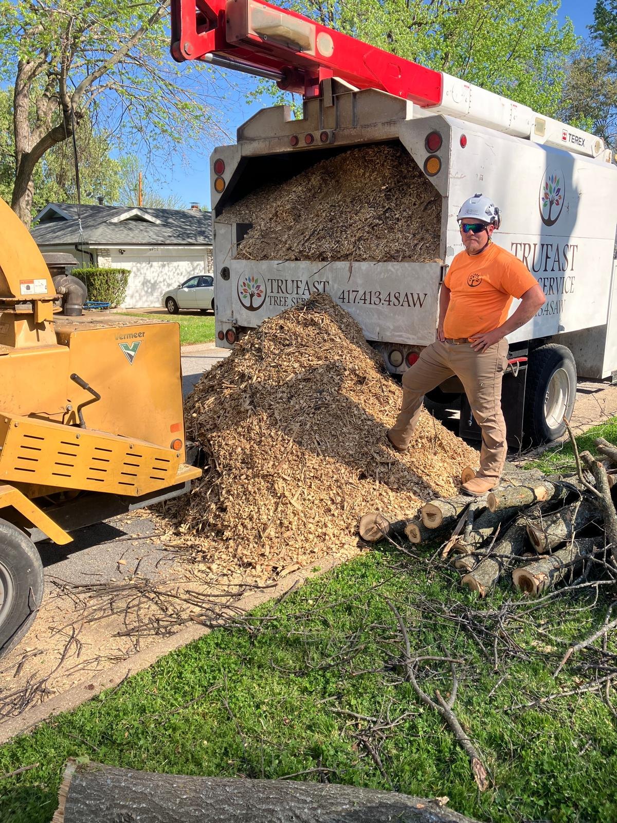 A Trufast crew member standing on a pile of fresh wood chips beside the chipper and the lettered chip truck.