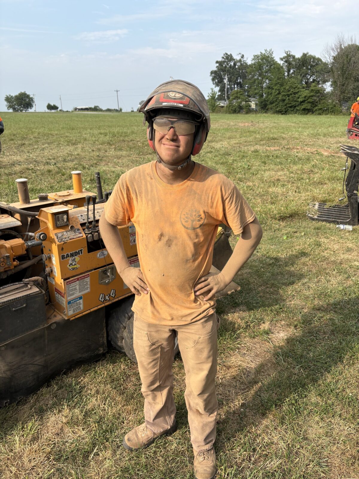 A grinning Trufast crew member covered head to toe in stump-grinding dust, hands on his hips beside the Bandit stump grinder.