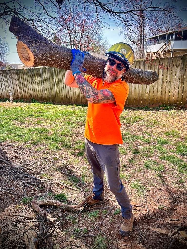 Doug Braudway, Trufast crew, grinning in a helmet and sunglasses with a big log over his shoulder