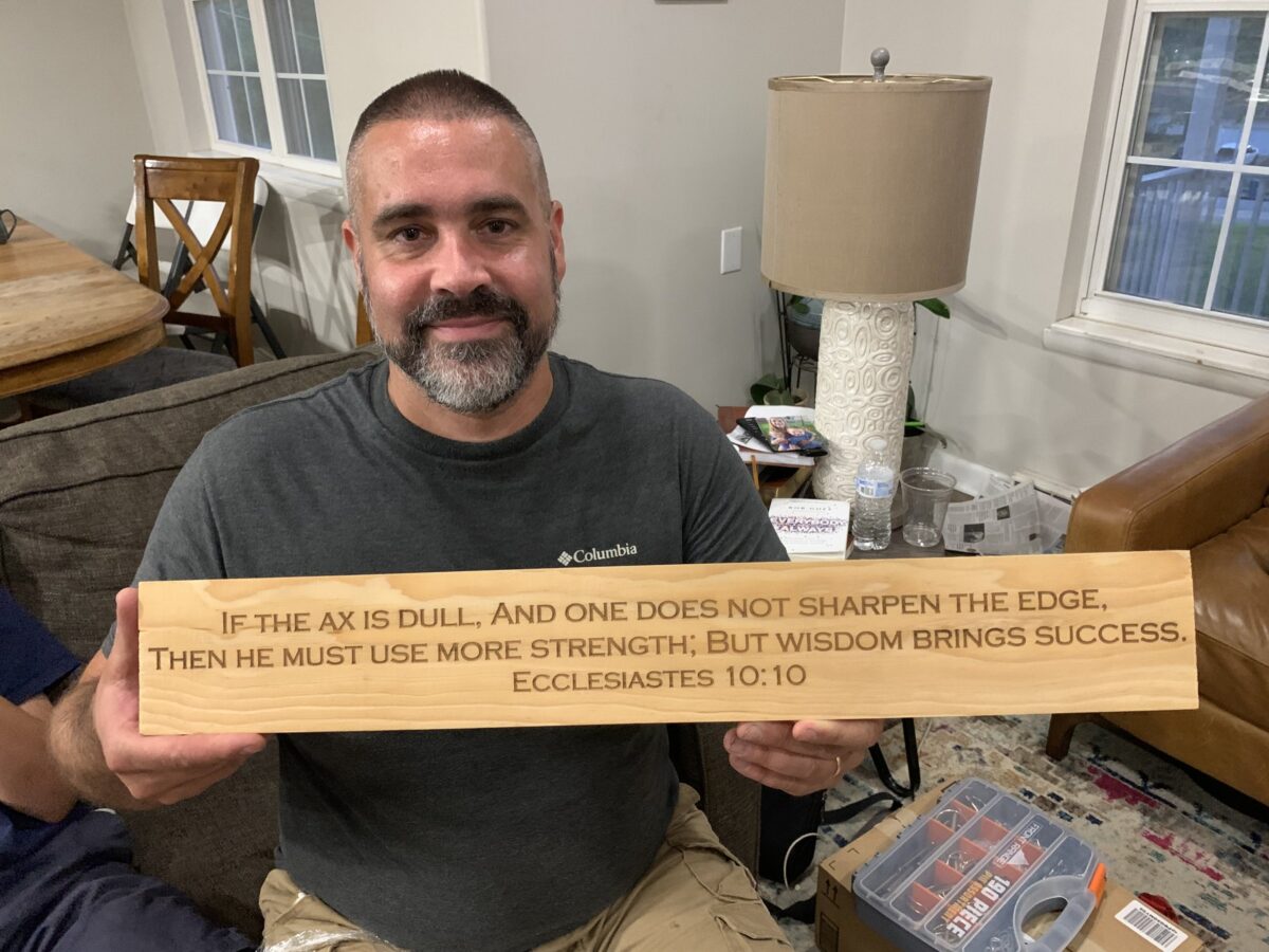A smiling man holding an engraved wooden sign that reads: If the ax is dull, and one does not sharpen the edge, then he must use more strength; but wisdom brings success. Ecclesiastes 10:10.