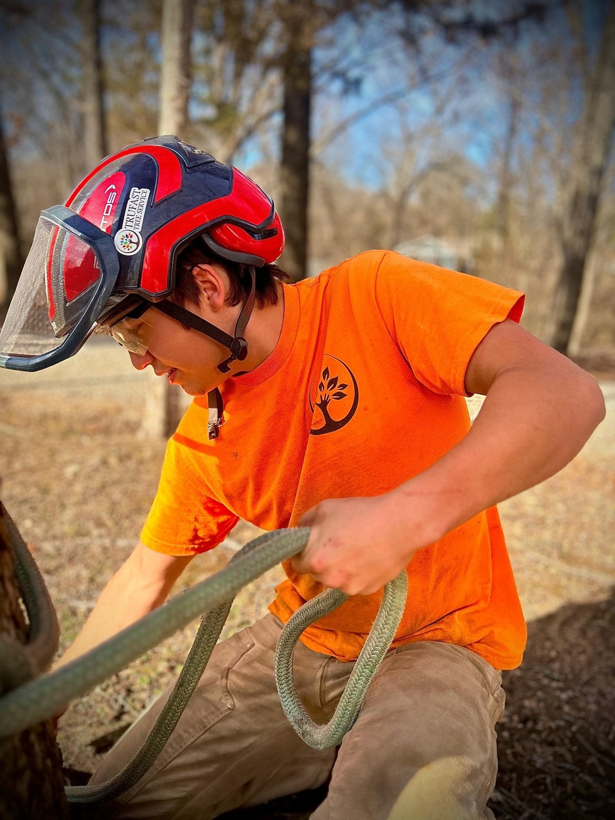 Josiah Bach, Trufast crew, in a red climbing helmet with face shield, handling a climbing rope at the base of a tree