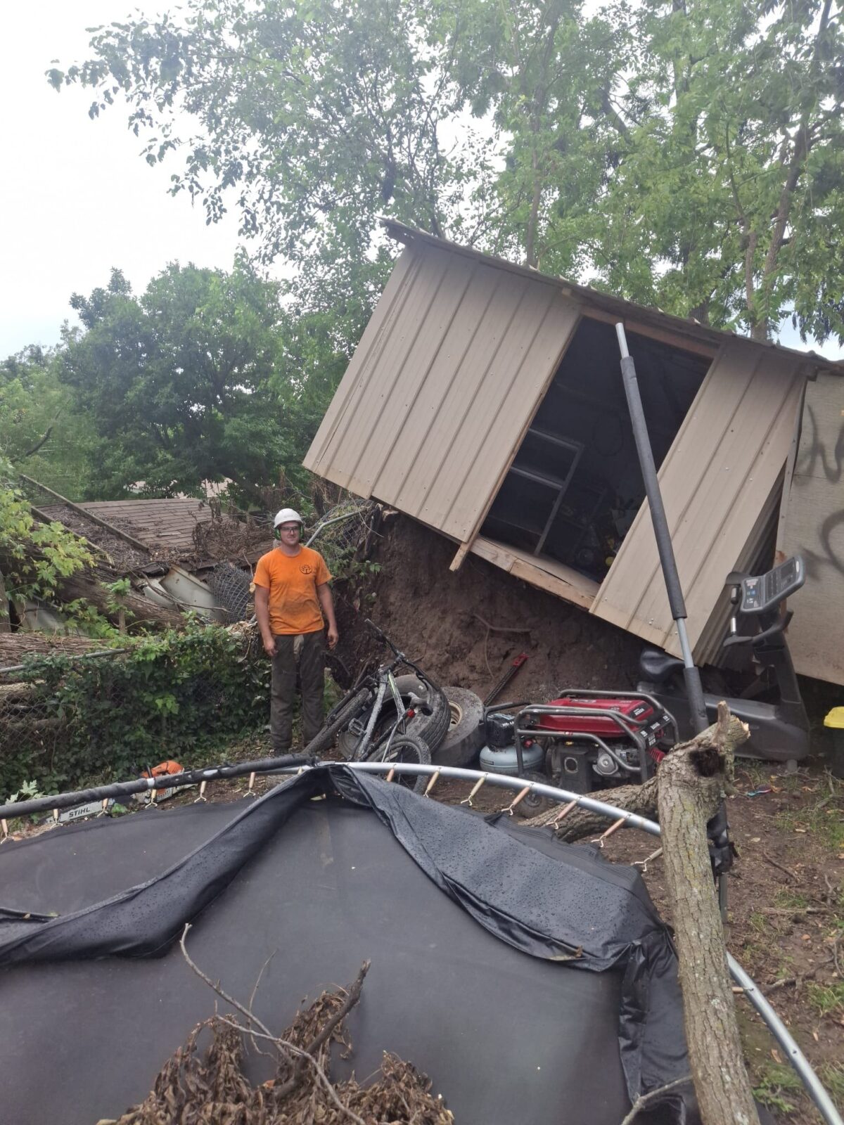 A storm-toppled tree whose root ball tore a shed off its foundation, a Trufast crew member beside it.