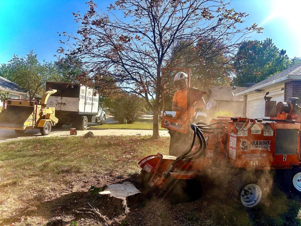 A Trufast crew member running a Bandit stump grinder into a fresh stump in a front yard, sawdust in the air, with the lettered Trufast truck and chipper parked behind.