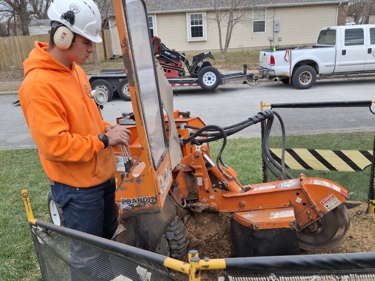 Trufast crew member in helmet and ear protection running a stump grinder behind a debris screen by the street