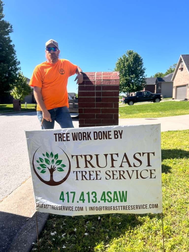 A Trufast crew member leaning on a brick mailbox beside a Tree Work Done By Trufast Tree Service yard sign in a neighborhood.