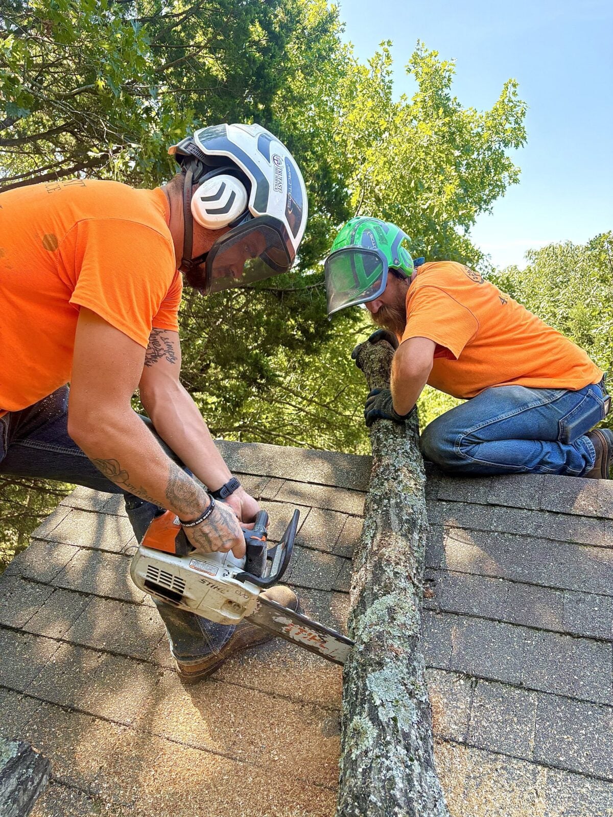 Two Trufast crew members in helmets and face shields on a roof, one cutting a limb with a chainsaw while the other holds it steady.